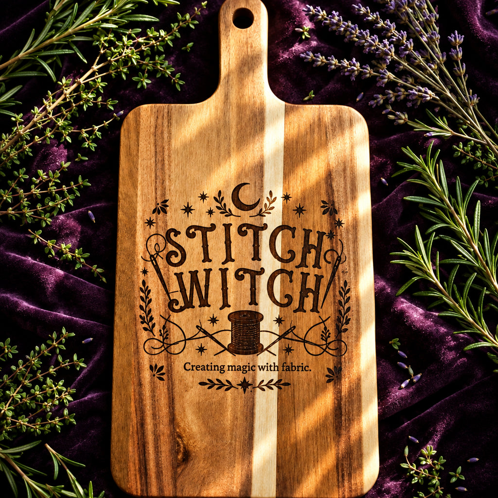 A wooden cutting board with a handle, featuring a decorative dark brown engraving that reads 'STITCH WITCH' with stars, a crescent moon, and a sewing machine. The board is placed on a wooden table with books visible in the background.