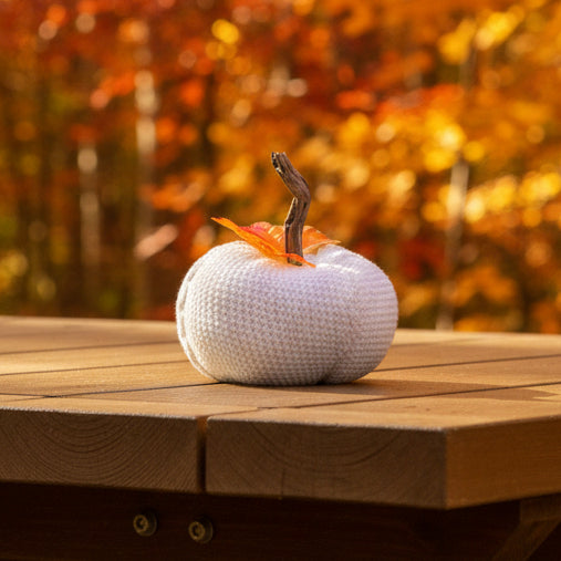 A white crocheted decorative pumpkin with a brown stem and orange-red autumn leaves, placed on a wooden deck outdoors.