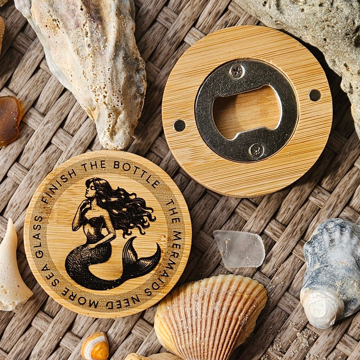 Two round wooden nautical-themed magnets and coasters with engraved mermaid designs, surrounded by various real beach shells and stones on a woven brown table background.