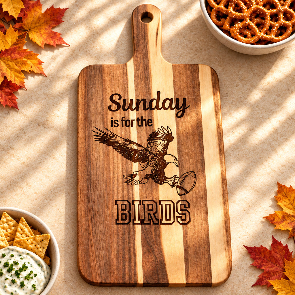 A wooden cutting board with a handle, featuring a natural wood grain pattern and a laser-engraved design that reads 'Sunday is for the BIRDS' with an eagle holding a coffee cup. The board is photographed on a white marble countertop.