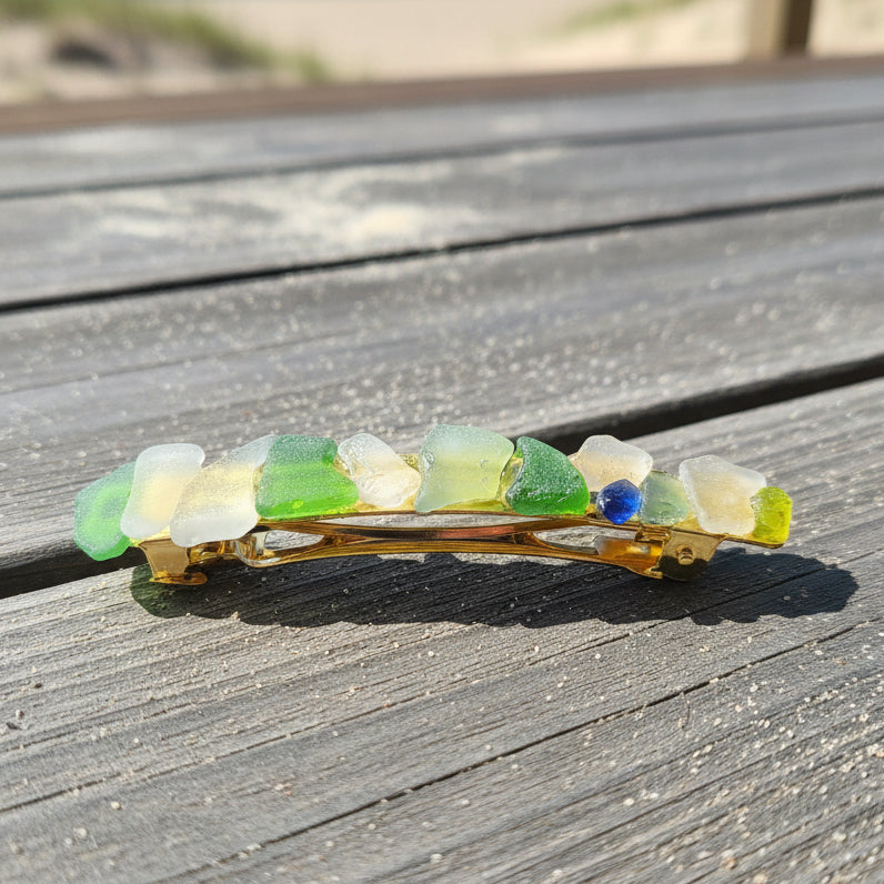 A small hair claw clip with a gold-tone metal body and a vertical row of translucent decorative sea glass in white, light green, and blue, resting on a brown wooden surface.