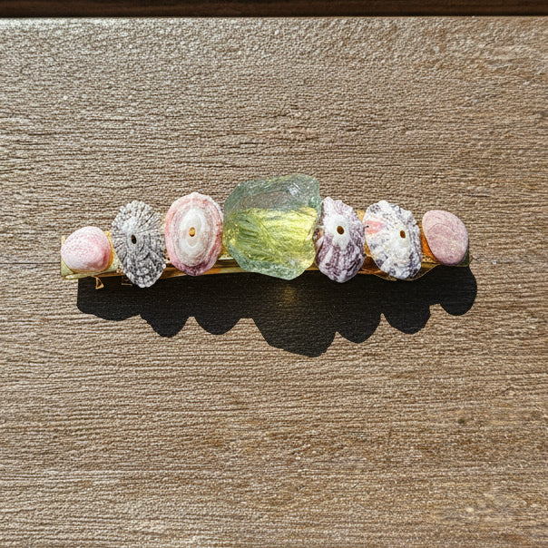 A gold-toned barrette hair clip decorated with pink seashells and a translucent green stone, resting on a brown wooden surface.
