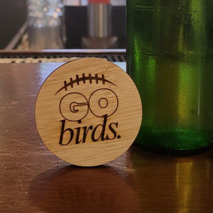A round wooden magnet featuring a football-shaped outline with the words 'GO birds' engraved, shown attached to a dark gray textured refrigerator door.