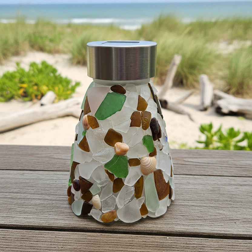 A sea glass mosaic solar light with a brushed stainless steel lid, sitting on a wooden deck with a blurred outdoor background of trees and grass.