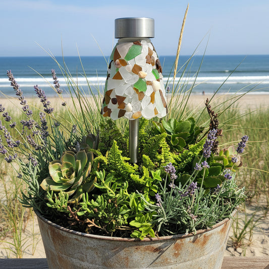 A mosaic solar garden light with a silver metal top and base, decorated with white, green, and brown sea glass pieces, shown outdoors in a planter box with green plants.