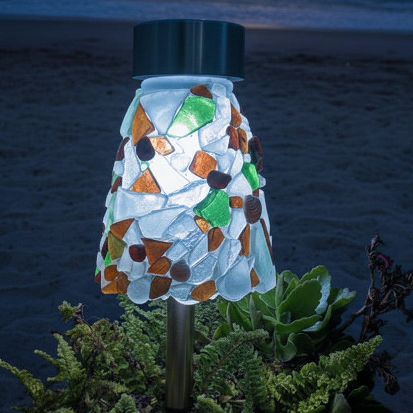A mosaic garden stake light with a conical shade made of white, green, and brown sea glass, planted in a galvanized metal pot filled with green ferns, set on a wooden deck outdoors.
