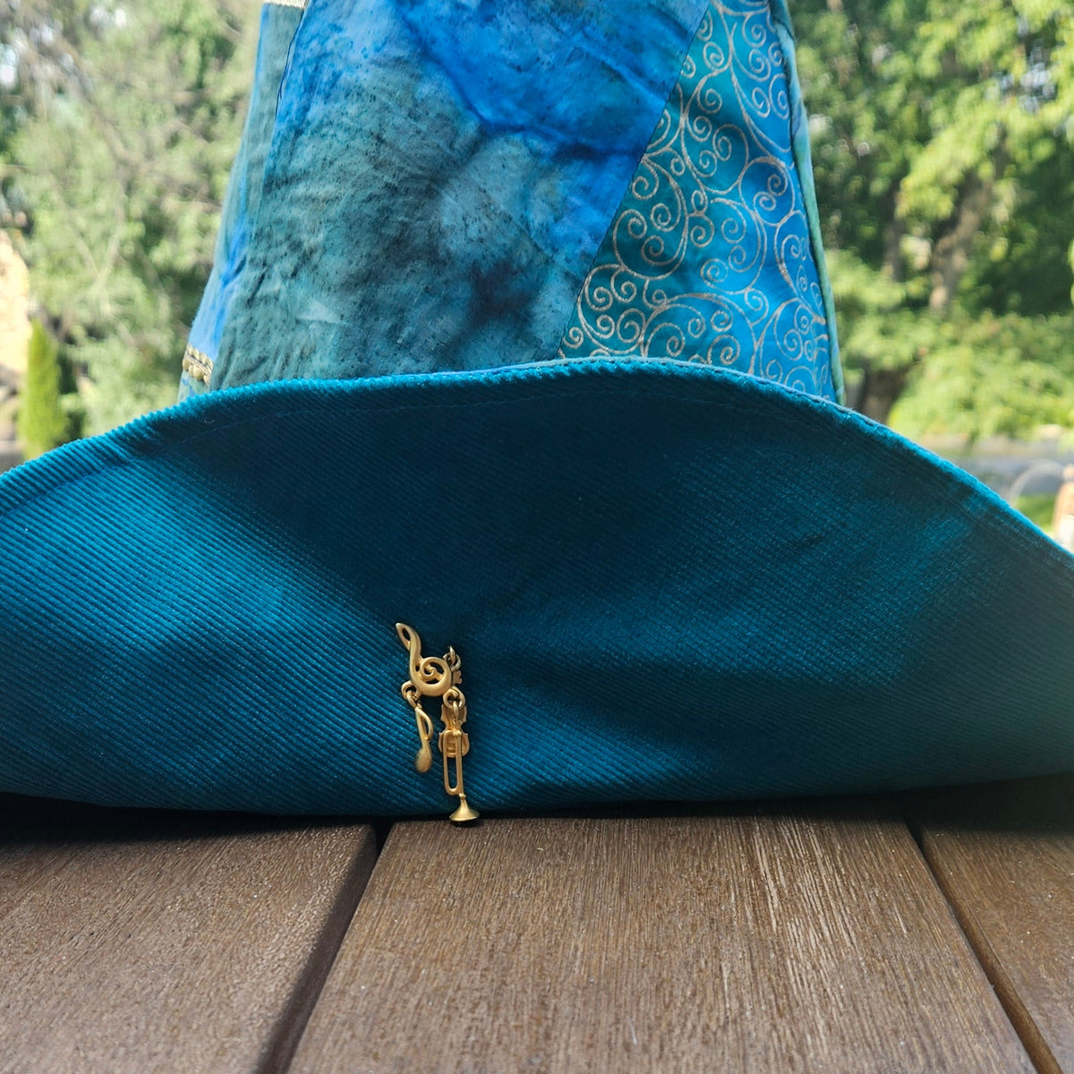 A blue and teal pointed witch hat with gold patterned trim and a small gold hanging charm, photographed outdoors on a wooden deck with trees in the background.