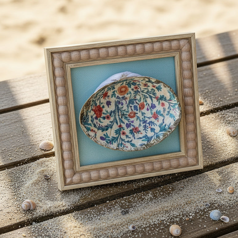 Decorative shell with floral design in a frame hanging on beachside table.