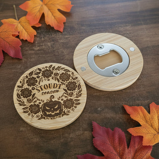A wooden magnet set on a dark wood tabletop, featuring a round light wood coaster with a metal bottle opener and a matching round coaster engraved with a floral wreath.